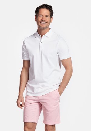 Man die lacht, draagt een wit polohemd met korte mouwen en lichtroze knielange korte broek, staat met één hand in de zak.