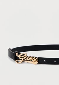 Μαύρη δερμάτινη ζώνη με αγκράφα από χρυσό μέταλλο με το λογότυπο "Guess", με λεία υφή και πολλαπλές τρύπες για ρύθμιση.