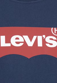 Tengerészkék póló, nagy piros Levi's logóval fehér felirattal, a háttérben jellegzetes íves mintával.