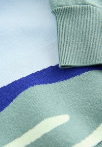 Nahaufnahme eines Strickpullovers mit Farbblocks in Hellblau, Grün, Marineblau und Creme sowie einem gerippten grünen Bündchen.