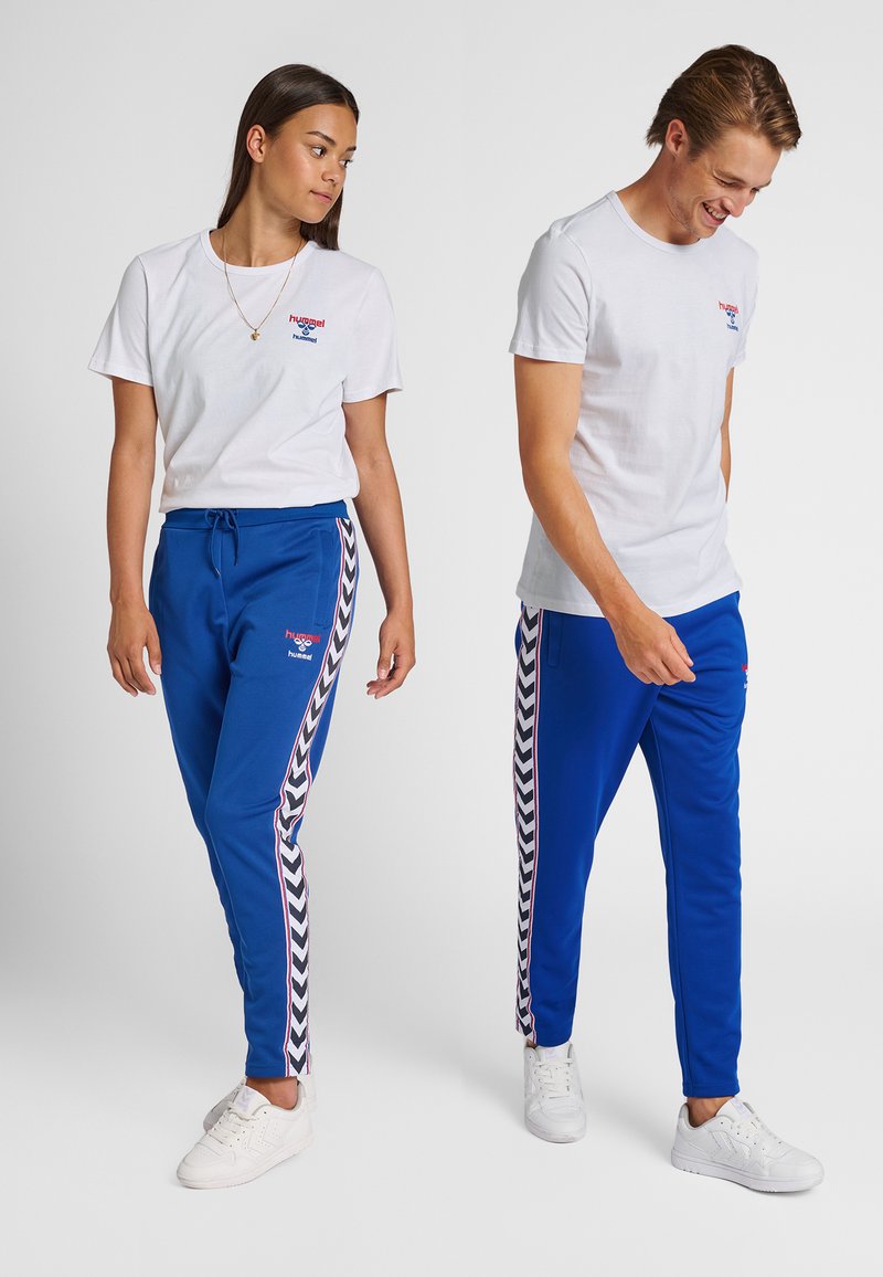 Ung kvinde og mand iført hvide T-shirts med logo og blå træningsbukser med sidelommer, stående mod en ensfarvet baggrund.