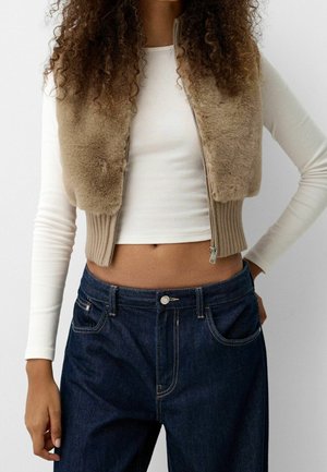 Mujer con chaleco sin mangas de piel sintética beige sobre una camiseta blanca de manga larga y corta, y jeans azul oscuro de talle alto con bolsillos.