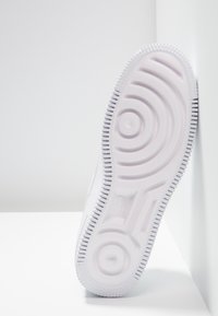 Semelle blanche de chaussure de sport montrant des motifs circulaires de la bande de roulement et des bords texturés, positionnée à la verticale contre un mur d’angle blanc.
