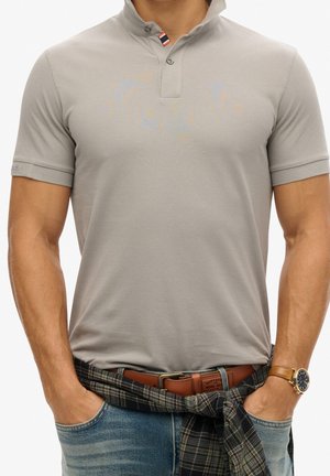 Man draagt een aangesloten lichtgrijs poloshirt, blauwe spijkerbroek, bruine riem, geruit overhemd om de taille gestrikt en een gouden horloge, handen in de zakken.