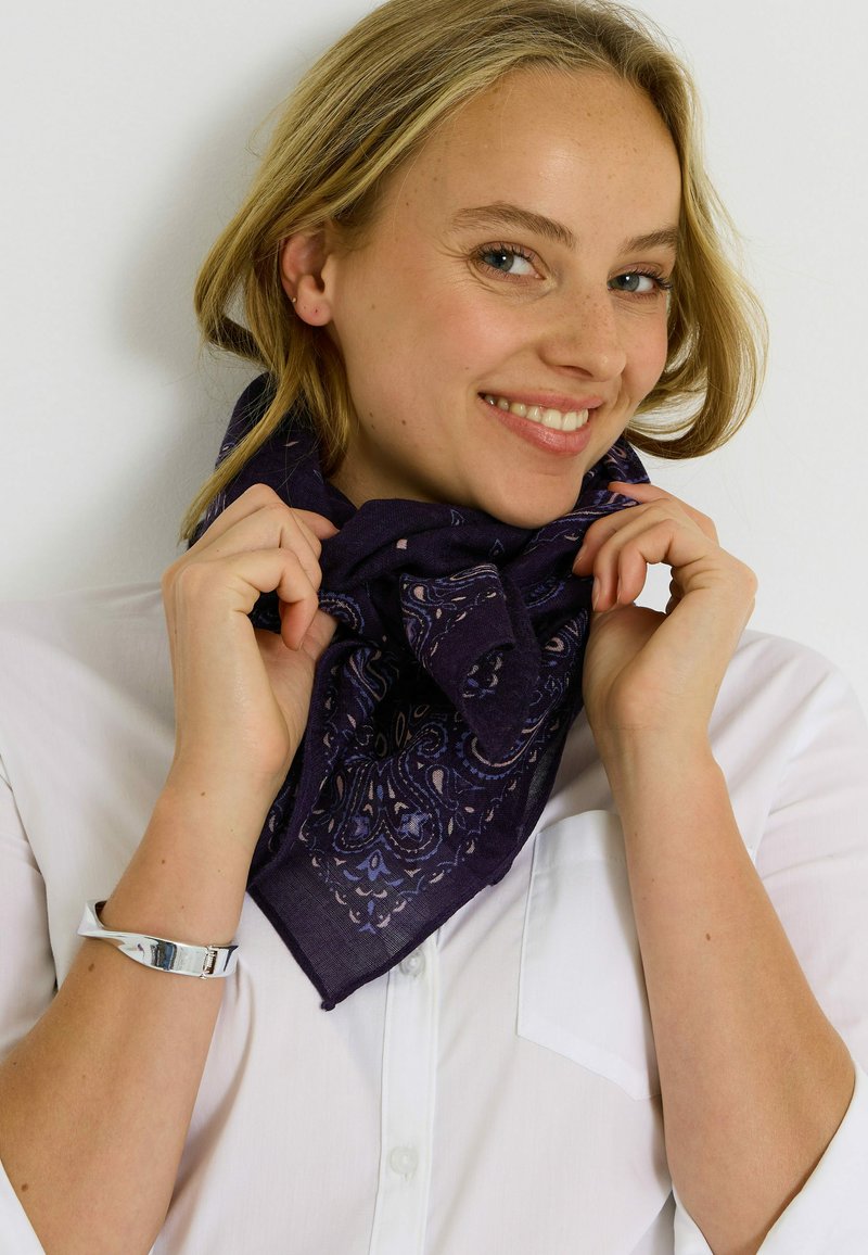 Bufanda morada oscura con patrón de paisley, llevada de manera suelta alrededor del cuello. Hecha de un tejido ligero, con una textura suave y bordes limpios.