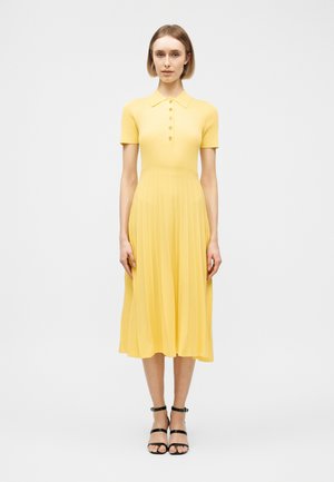 Γυναίκα που στέκεται φορόντας ένα κίτρινο midi φόρεμα με κοντά μανίκια, κουμπωτό και πλισέ φούστα, μαζί με μαύρα σανδάλια με λουράκια σε λευκό φόντο.