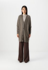 Cardigan lung, supradimensionat, dintr-un material moale, purpuriu, cu decolteu în V, închidere cu șase nasturi și mâneci largi. Asortat cu pantaloni largi, negri.