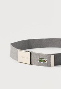 Ceinture en tissu gris avec boucle métallique argentée gravée "Lacoste" et logo crocodile vert brodé à l'extrémité de la sangle.