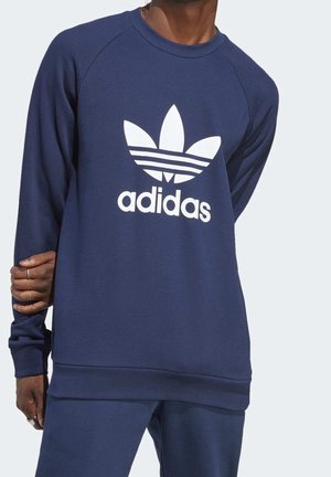 Mand iført marineblå Adidas-sweatshirt og matchende bukser, med hvidt Adidas trefoil-logo centreret på sweatshirten.