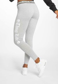 Grijze leggings met zwarte tekst "Thug Life" en een schedelafbeelding aan de zijkant. Voorzien van een zwarte elastische tailleband. Gecombineerd met witte sneakers.