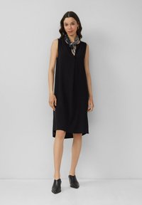 Schwarzes ärmelloses Kleid mit V-Ausschnitt, drapiertem Design und einem Saum, der unter dem Knie endet; accessoriert mit einem gemusterten Halstuch.