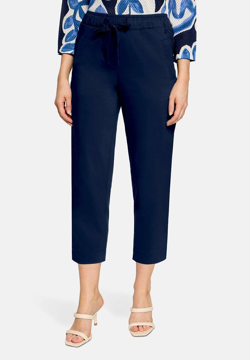 Femme portant un pantalon court bleu marine avec taille à cordon et des sandales à talons beiges, debout sur un fond uni.