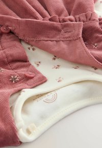 Roze corduroy top met knoopsluiting en ruffle-details, gelaagd over een crèmekleurige stof met een klein bloemenpatroon. Zachte textuur, casual ontwerp.