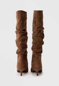Botas de ante marrón, de caña alta y caídas, con tacones de media altura, vistas desde atrás sobre un fondo blanco liso.