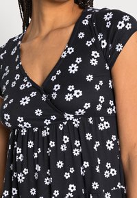Musta mekko, jossa on V-pääntie ja lyhyet hihat. Tässä mekossa on valkoisten päivänkakkaroiden kukkakuosi ja rypistetty vyötärö, joka luo virtaavan siluetin.
