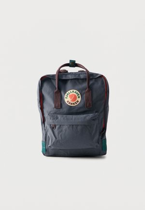 Rucsac Fjällräven Kånken albastru închis, cu buzunar frontal cu fermoar, mânere maro și patch rotund cu logo pe un fundal alb simplu.