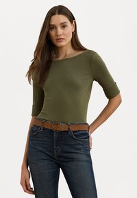 Olijfgroene aansluitende top met een wijde halslijn en driekwart mouwen, gecombineerd met donkerblauwe jeans en een bruine riem. Soepele, rekbare stof.