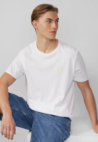 Weiße Baumwoll-T-Shirt mit rundem Halsausschnitt, kurzen Ärmeln und einem dezenten gelben Grafikdesign auf der linken Brust. Lässige Passform.