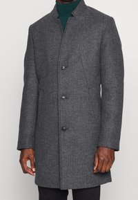 Cappotto herringbone grigio dal design strutturato, con colletto alto, cinque bottoni neri e taglio dritto. Adatto per abbigliamento formale.