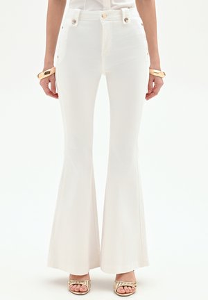 Pantalon évasé blanc taille haute avec des boutons latéraux, porté avec des bracelets dorés et des chaussures à talons ouverts à rayures métalliques.