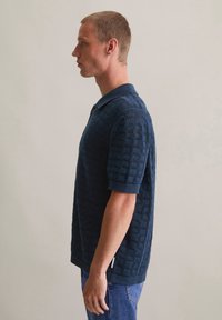 Donkerblauw poloshirt met een gestructureerd geometrisch patroon, korte mouwen en een kraag, gecombineerd met denim jeans.