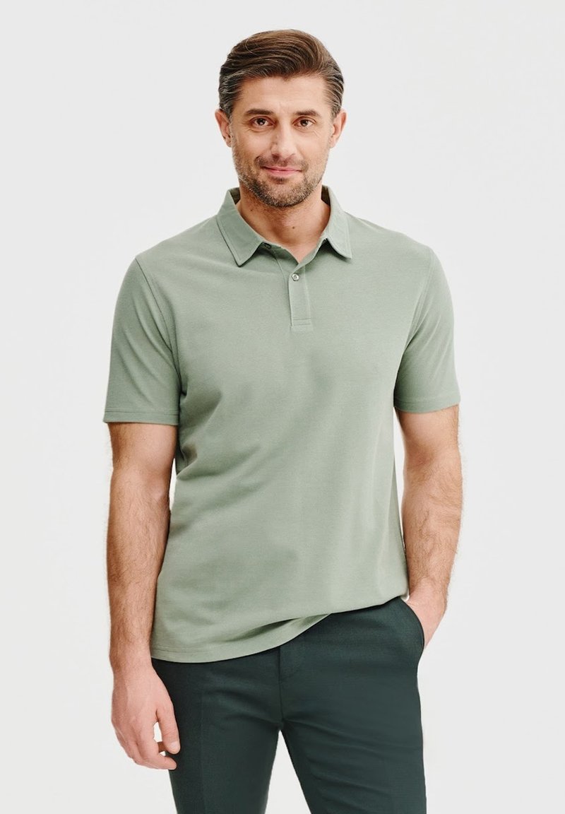 Koszulka polo z krótkim rękawem w miękkim zielonym kolorze, z klasycznym kołnierzykiem, dwoma guzikami i luźnym krojem. Noszona z ciemnymi spodniami.