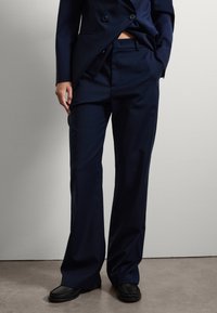 Pantaloni navy realizzati in un tessuto liscio, con una vestibilità sagomata e gamba dritta, pieghe sottili e tasche laterali. Scarpe nere slip-on completano il look.