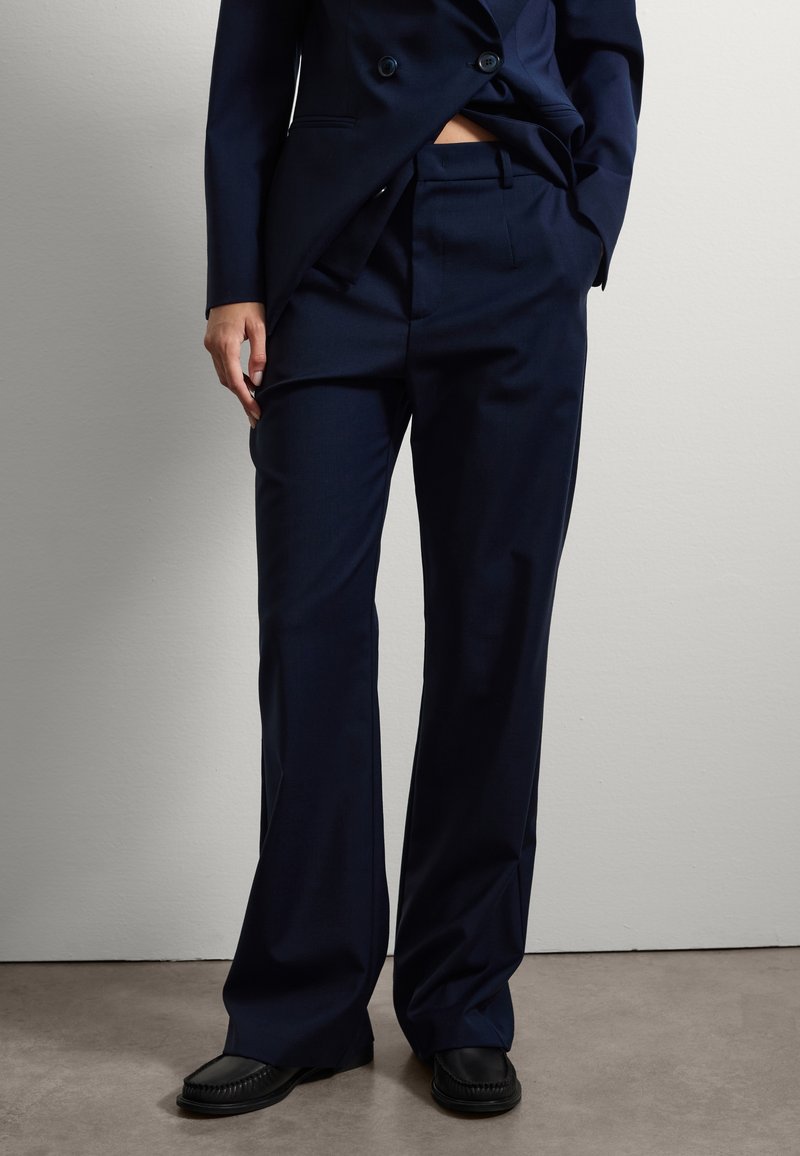 Pantaloni navy realizzati in un tessuto liscio, con una vestibilità sagomata e gamba dritta, pieghe sottili e tasche laterali. Scarpe nere slip-on completano il look.