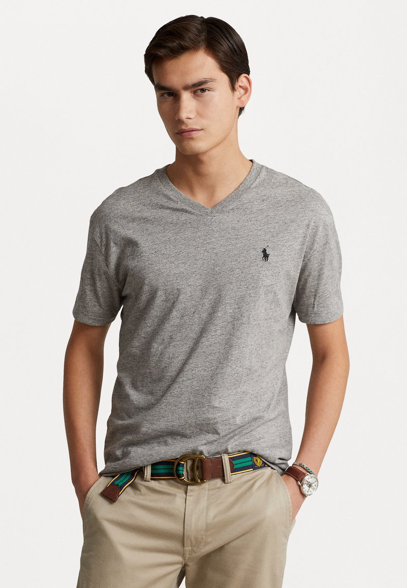 Camiseta gris de cuello en V hecha de una tela texturizada, con mangas cortas y un pequeño logo en navy. Combinada con pantalones beige y un cinturón estampado.