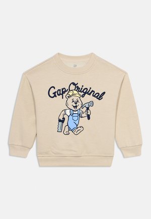 Beiger Kinderpullover mit einem Cartoon-Bären in blauer Latzhose, der einen Hammer und eine Säge hält, mit dem Text "Gap Original" darüber.