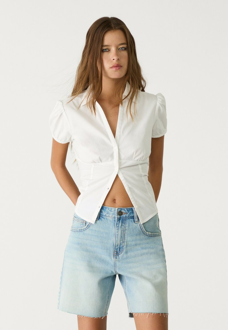 Bela kratka majica s kratkimi rokavi in detajli gumbov ter nabranimi rameni, v kombinaciji s svetlo modrimi visokimi jeans kratkimi hlačami z raztrganim robom.