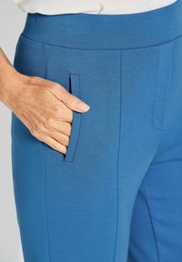 Blaue Hose mit einer glatten Textur, die einen horizontalen Bund und eine Tasche an der Seite hat. Die Hand liegt in der Tasche.