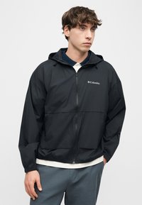 Νέος άνδρας που φοράει μαύρο φούτερ Columbia με φερμουάρ και κουκούλα πάνω από λευκό πουκάμισο, τα χέρια στις τσέπες, κοιτάζοντας πλάγια μπροστά σε απλό φόντο.