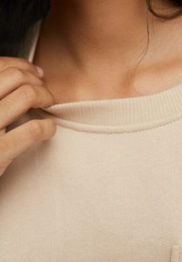 Beige Baumwollpullover mit einem gerippten Kragen. Nahaufnahme zeigt eine glatte Textur und eine Hand, die sanft am Halsausschnitt zieht.