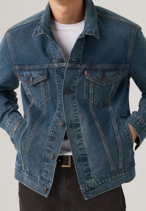 Personne portant une veste en jean bleue par-dessus une chemise blanche avec les mains dans les poches latérales, boucle de ceinture visible et pantalon foncé.