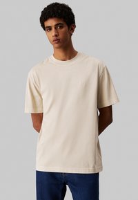 T-shirt beige a maniche corte in cotone con scollatura rotonda e logo ricamato discreto sul lato sinistro del petto. Tessuto morbido, vestibilità rilassata.