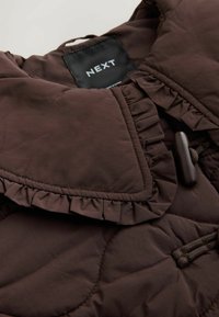 Veste matelassée marron foncé avec bords à volants, fermeture par bouton à bascule et label de marque NEXT visible à l’intérieur du col.