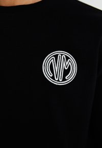 Svart sweatshirt med et sirkulært hvitt brodert logo med stiliserte bokstaver, satt mot en teksturert stoffbakgrunn.