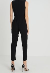 Tutina nera senza maniche con vita aderente, design a gamba affusolata e texture liscia. Abbinata a scarpe nere con tacco alto.