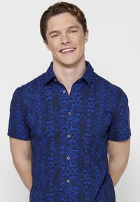 La camicia a maniche corte presenta un motivo astratto blu e blu scuro con bottoni beige, realizzata in tessuto leggero e con un design di colletto classico.