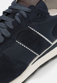 Jūras spēku sporta apavi ar zamša un tekstila paneļiem, kontrastējošām baltajām svītrām, teksturētām šņorēm un diskrētu brūnu akcentu uz mēles.