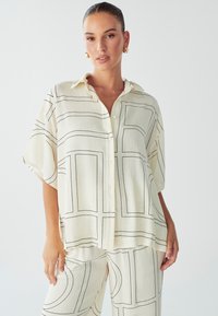 Camisa blanca de botón con líneas rectangulares negras y una textura suave. Cuenta con un cuello y un corte holgado, combinada con pantalones de patrón a juego.