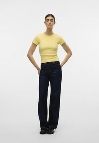 T-shirt gialla aderente e jeans dritti blu scuro. La t-shirt ha maniche corte; i jeans presentano uno stile classico a cinque tasche e accessori in metallo.