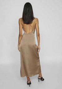 Bruine satijnen maxidress met een lage rug en dunne schouderbandjes, met een gladde textuur en een zijsplit. Zwarte sandalen met bandjes complementeren de look.