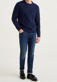 Man som bär en marinblå mönstrad stickad tröja, mörkblå jeans och svarta tofflor med fransar, med en hand i fickan mot en enfärgad bakgrund.
