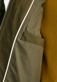 Jachetă verde măslinie cu fermoar alb, dintr-un material texturat și cu un buzunar lateral. Fundalul prezintă un material cu model în fagure de miere de culoare muștar.