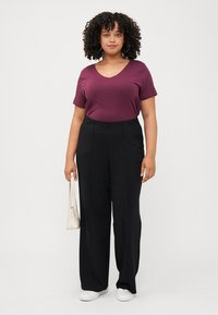 Woman with curly hair wears a maroon v-neck shirt and black wide-leg trousers, holding a white bag, standing against a white background.