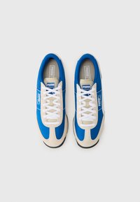 Sneaker blu e beige con tomaia in tessuto liscio, lacci bianchi e suola strutturata. Include un logo sul lato e un collo imbottito.