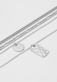 Des colliers en argent superposés avec un pendentif rectangulaire texturé et un charm circulaire lisse, le tout sur des chaînes délicates de styles variés.