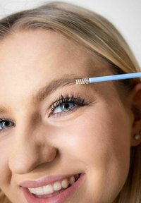 Blå øyenbrynspensel med spiralmønstrete bust holdt nær en kvinnes bryn, som viser hennes lange vipper og lyse hår. Huden har en jevn tekstur.